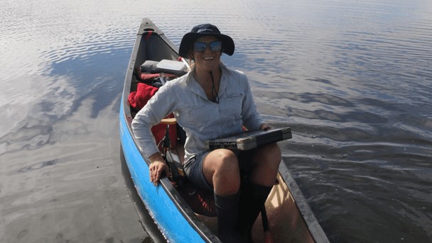 A woman sits in a canoe on a calm waterway. She is holding a clipboard and wearing a hat, outdoor clothing and gumboots.
