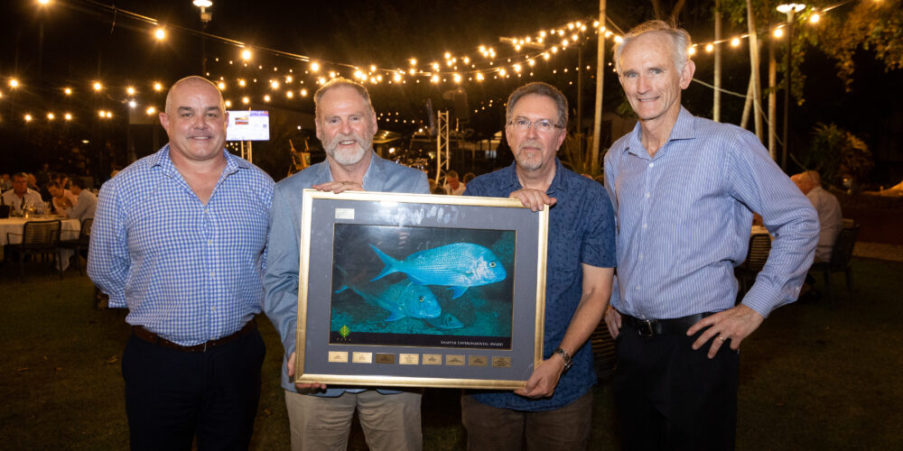 Four men stand in a row holding a large frame with a picture of two blue fish. They are standing outside at night with festoon lighting in the background.